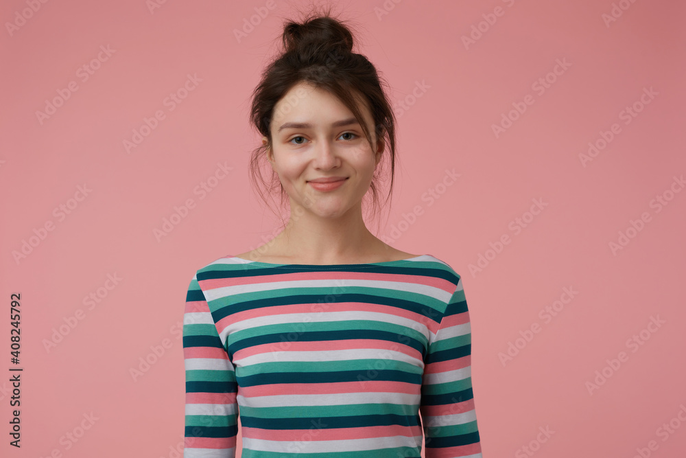 Young lady, pretty brunette woman with bun. Wearing striped blouse and have cute smile, happy to see you, showing confidence. Watching at the camera isolated over pastel pink background
