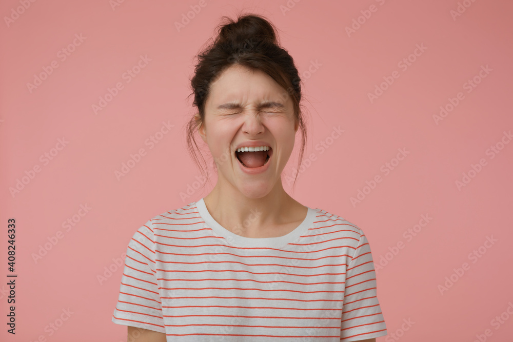 Teenage girl, angry looking woman with brunette hair and bun. Wearing t-shirt with red strips and yelling in anger. Emotional concept. Stand isolated over pastel pink background Teenage girl, angry looking woman with brunette hair and bun. Wearing t-shirt with red strips and yelling in anger. Emotional concept. Stand isolated over pastel pink background