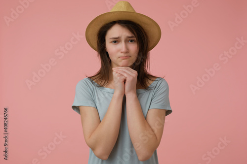 Teenage girl, woman with begging look and long brunette hair. Wearing blueish t-shirt and hat. Holding hands under her chin and ask. Watching at the camera isolated over pastel pink background