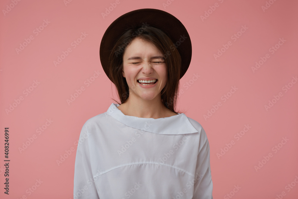 Teenage girl, happy looking woman with long brunette hair. Wearing white blouse and black hat. Laughing and keep eyes closed. Emotional concept. Stand isolated over pastel pink background Teenage girl, happy looking woman with long brunette hair. Wearing white blouse and black hat. Laughing and keep eyes closed. Emotional concept. Stand isolated over pastel pink background