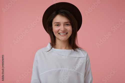 Portrait of attractive, happy girl with long brunette hair. Wearing white blouse and black hat. Having a wide smile. Emotional concept. Watching at the camera isolated over pastel pink background © timtimphoto