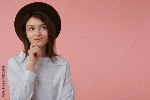 Wonder looking woman, beautiful girl with long brunette hair. Wearing white blouse and black hat. Touch her chin. Emotional concept. Watching to the right at copy space over pastel pink background