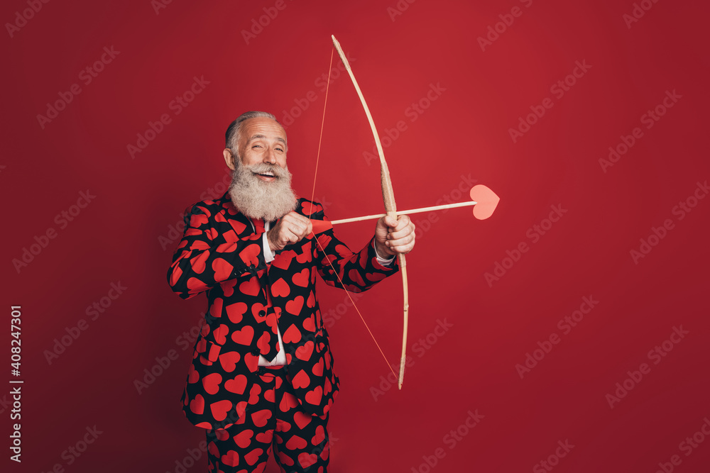 Photo of positive retired cherub man hold bow arrow prepare shoot wear heart print tux isolated red background - obrazy, fototapety, plakaty 