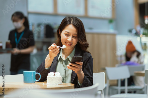 Portrait of young Asian businesswoman drinking coffee  eating cream cake and using phone
