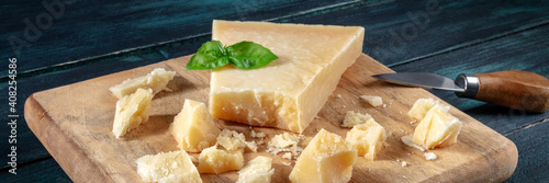 Parmesan cheese panorama with crumbles, basil leaves and a cheese knife on a dark blue wooden background