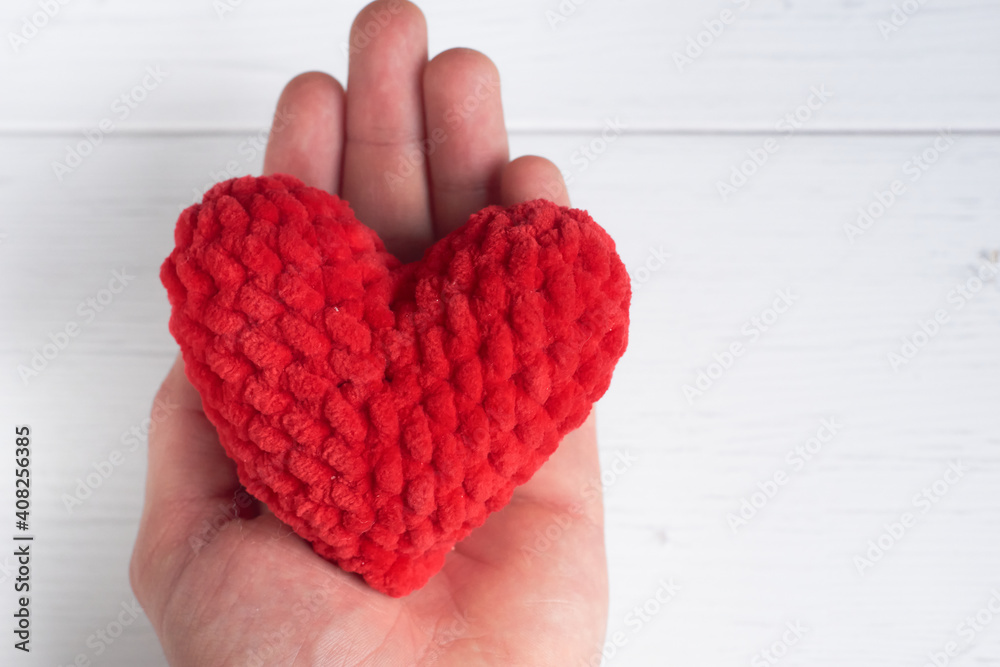 Male Caucasian hand holding a red knitted heart on an old white wooden background with copy space for text. The concept of love, valentine's day, care. Selective focus. Male Caucasian hand holding a red knitted heart on an old white wooden background with copy space for text. The concept of love, valentine's day, care. Selective focus.