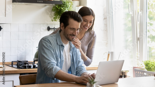 Sharing good news. Happy smiling millennial man use laptop at home office on kitchen typing pleasant message. Caring young woman wife standing by hugging husband with warmth reading email from screen