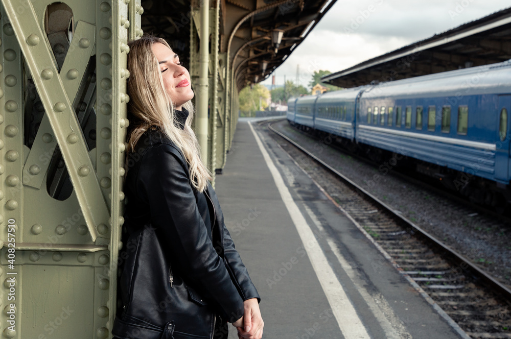 Portrait of a young beautiful blonde in a leather jacket with closed eyes on the platform of the railway station.