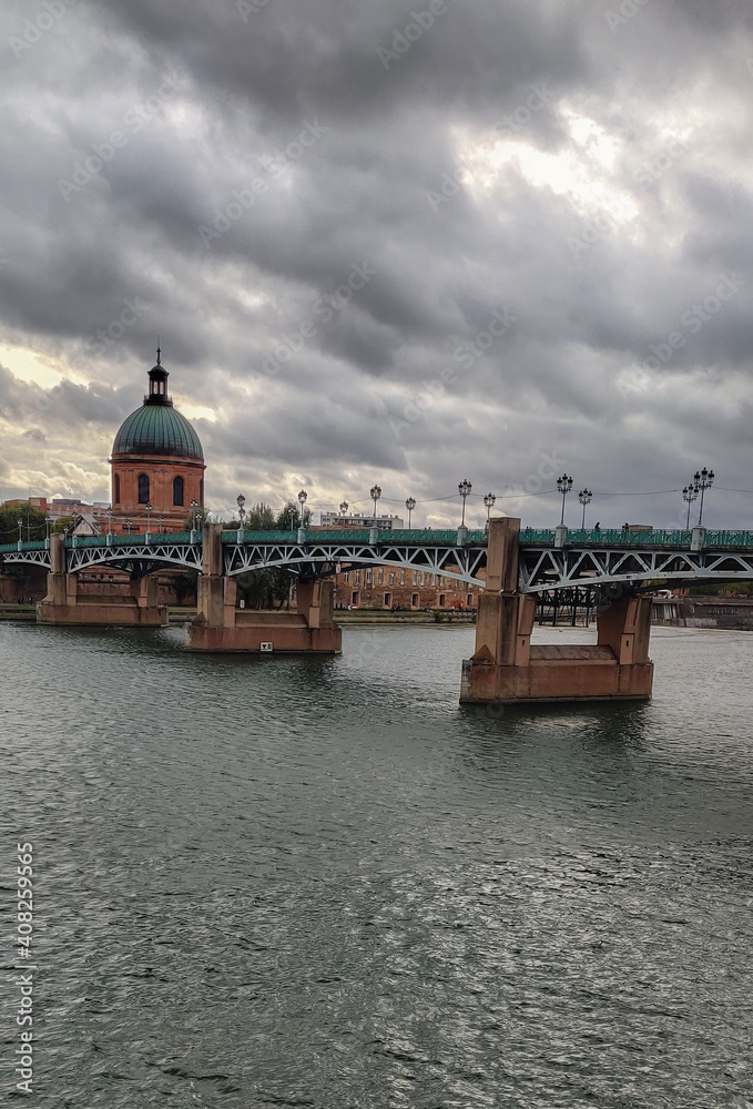 130.	Brick and metallic bridge with lanterns crossing the river. Dome of a historic building on the background. Cloudy sky and rainy weather. Toulouse, France