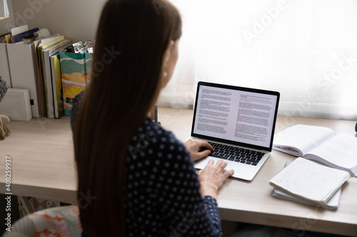 Paperwork on computer. Back view of young lady working from home focused on pc screen read electronic book. Millennial woman study digital document at online database apply corrections to draft report