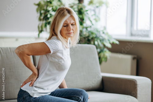 Photos Upset mature woman suffering from backache, unhappy senior blonde sitting on a sofa at living room, feeling discomfort because of pain in back