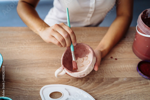 Close-up of girl painting clay mug. Woman coloring pottery in workshop with a paintbrush. Painter in white tanktop enthusiastically glazing cup.