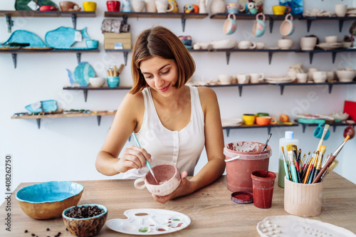 Mid-shot of potter decorating clay mug after firing in oven. Woman in white tanktop enopying creative procces of pottery coloring. Sitting in workshop with white walls and lorful clay items, products