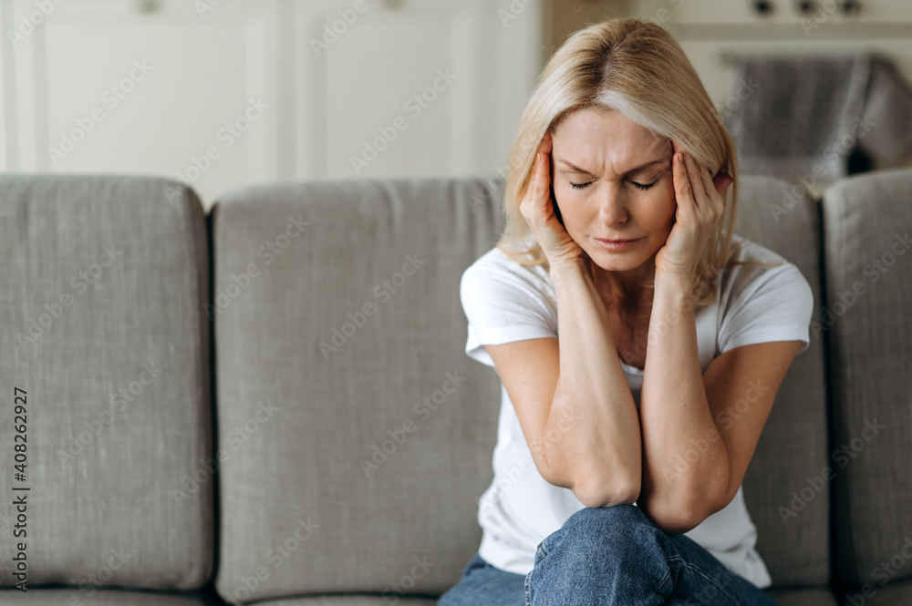 Middle aged blonde woman sits on couch at living room holding her head with her hands, feels unhappy because of headache, personal troubles, illness or bad news, she need psychological or medical Middle aged blonde woman sits on couch at living room holding her head with her hands, feels unhappy because of headache, personal troubles, illness or bad news, she need psychological or medical