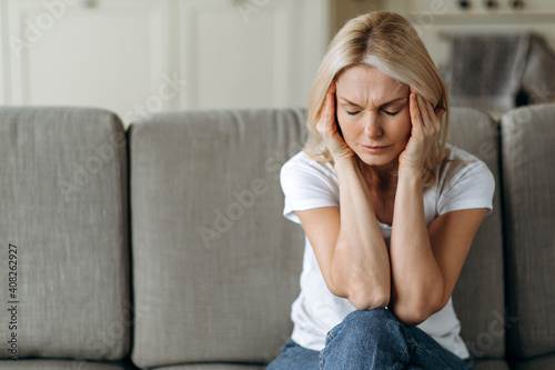 Middle aged blonde woman sits on couch at living room holding her head with her hands, feels unhappy because of headache, personal troubles, illness or bad news, she need psychological or medical