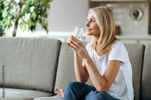 Healthcare concept. Mature caucasian blonde dressed in a casual wear sits on the couch in a living room, holding a glass of pure water in a hand and smiling, follow healthy lifestyle