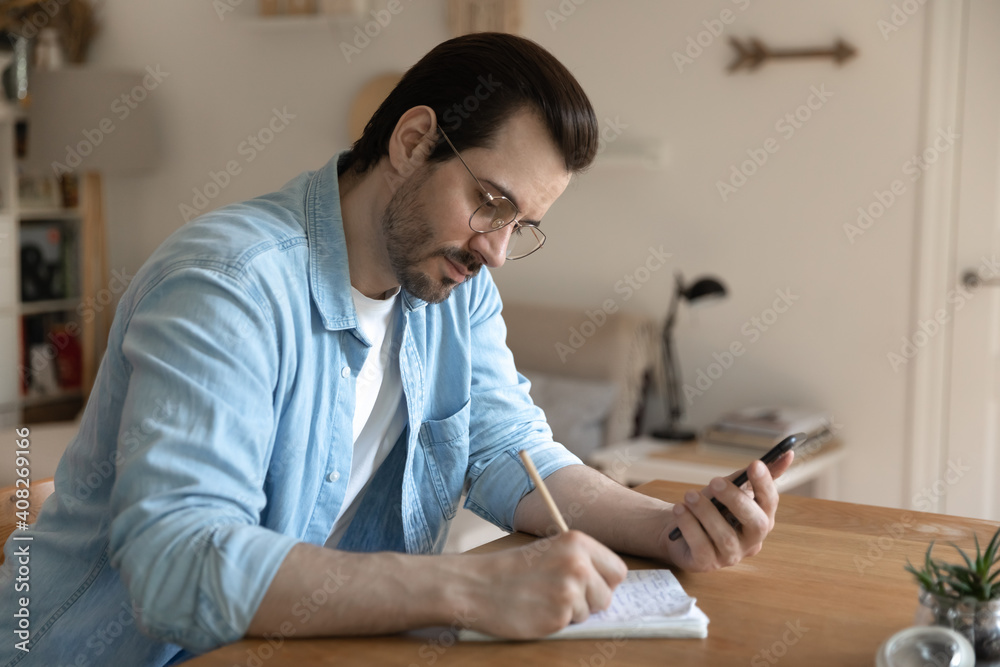 Focused young male student sitting by table holding smartphone listening to distant lecture making brief notes. Businessman freelancer watching training course writing up details to paper workbook Focused young male student sitting by table holding smartphone listening to distant lecture making brief notes. Businessman freelancer watching training course writing up details to paper workbook