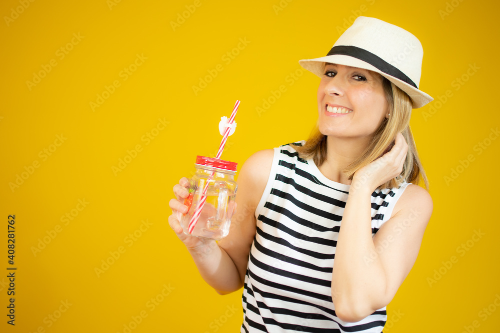Smiling woman wearing straw hat holding orange juice glass over yellow background. Smiling woman wearing straw hat holding orange juice glass over yellow background.