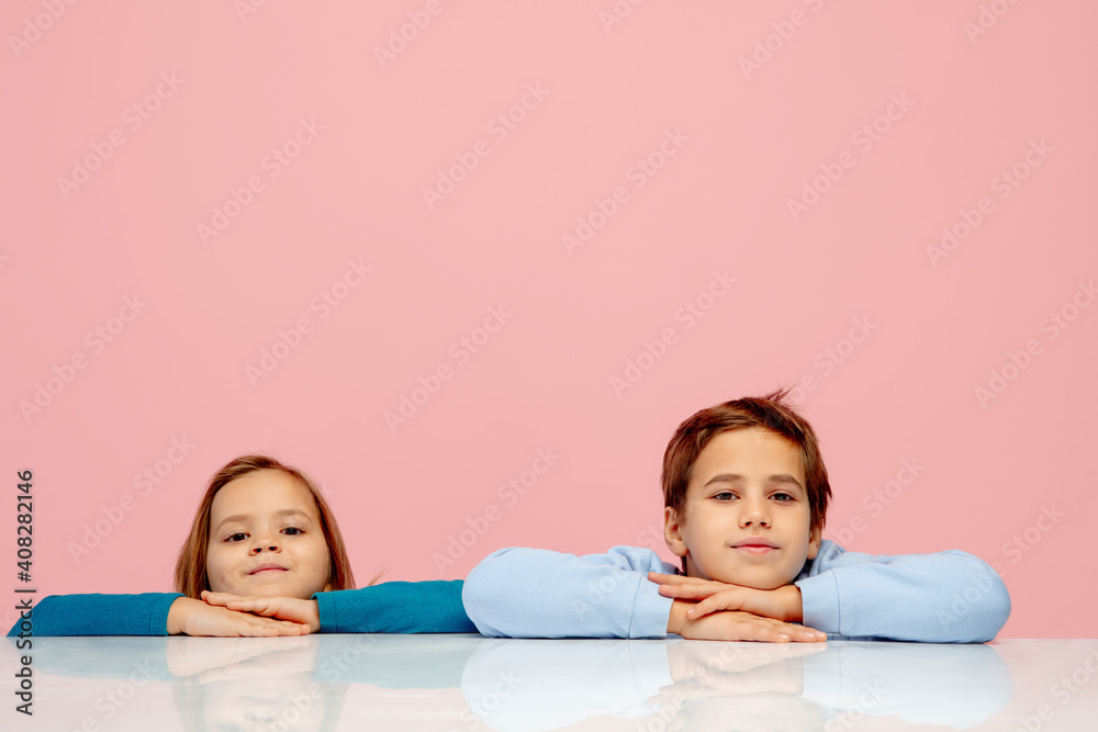 Best friends. Happy children isolated on coral pink studio background. Look happy, cheerful. Copyspace for ad. Childhood, education, emotions, facial expression concept. Sitting at the table, school Best friends. Happy children isolated on coral pink studio background. Look happy, cheerful. Copyspace for ad. Childhood, education, emotions, facial expression concept. Sitting at the table, school