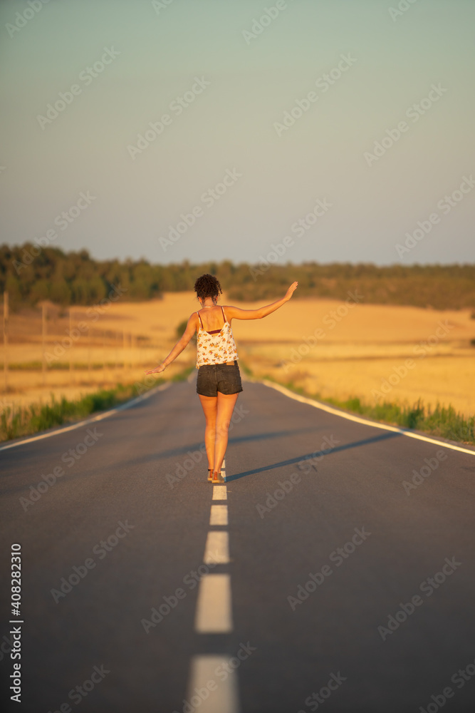 Young girl, walking in the middle of a lonely straight road, late in the day. With the hands stretched out in the shape of a cross, to maintain balance and not fall from the dividing line.