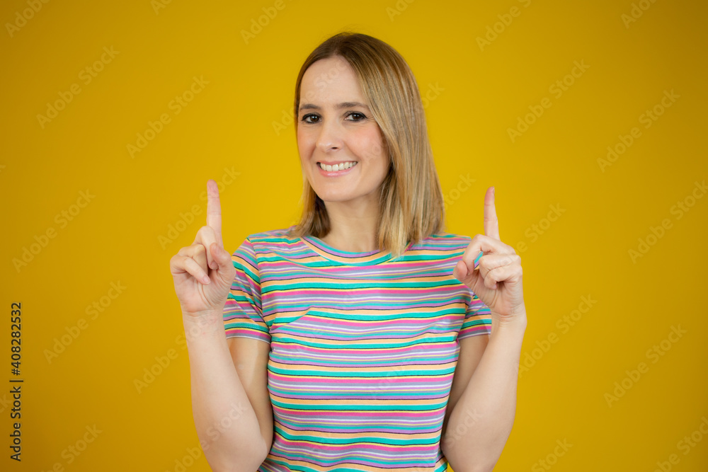 Young woman pointing up with both index fingers while smiling cheerfully at camera, standing over yellow background. Young woman pointing up with both index fingers while smiling cheerfully at camera, standing over yellow background.