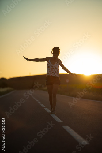 Fototapeta Naklejka Na Ścianę i Meble -  Young girl, walking in the middle of a lonely straight road, late in the day. With the hands stretched out in the shape of a cross, to maintain balance and not fall from the dividing line.