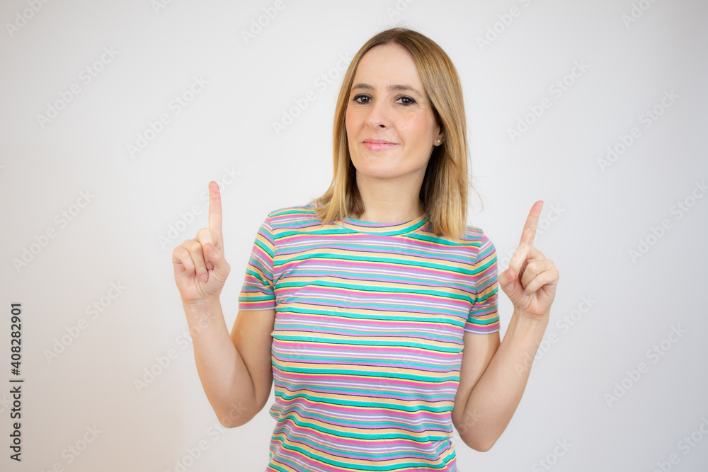 Young woman pointing up with both index fingers while smiling cheerfully at camera, standing over white background.