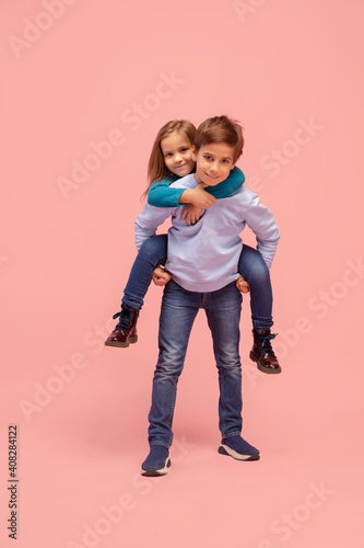 Togetherness. Happy children isolated on coral pink studio background. Look happy, cheerful. Copyspace for ad. Childhood, education, emotions, facial expression concept. Having fun, piggyback riding