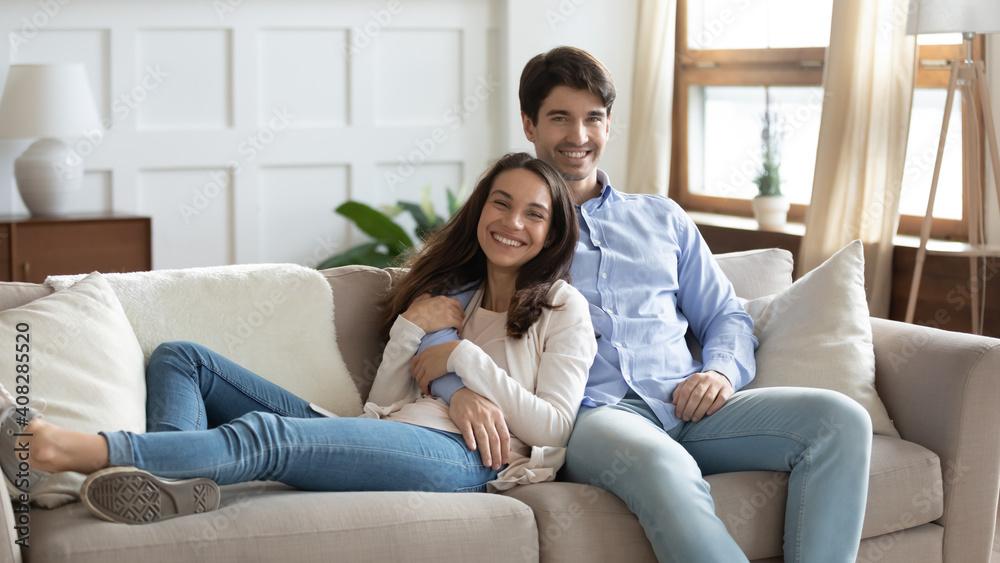 Family portrait smiling woman and man hugging, sitting on cozy couch in living room at home, happy young couple posing for photo, loving wife and husband cuddling, looking at camera together