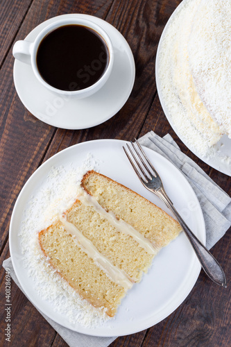 Piece of homemade coconut cake with cream cheese frosting and coconut flakes decoration, on a white plate, vertical, top view, closeup