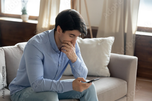 Close up thoughtful serious man touching chin, looking at smartphone screen, having problem with phone or reading bad news in message, unexpected debt notification, upset male sitting on couch