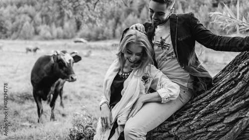 Handsome guy and blonde girl walking on the grass near the river and forest. They hold hands and smile on the background of pasture with cows and horses