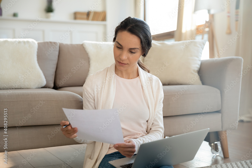 Close up serious young woman reading letter, holding paper sheet, sitting on warm floor with laptop on laps, focused businesswoman working with correspondence, checking documents at home Close up serious young woman reading letter, holding paper sheet, sitting on warm floor with laptop on laps, focused businesswoman working with correspondence, checking documents at home