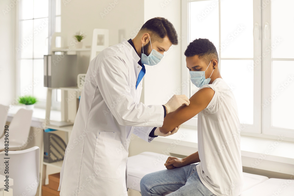 Doctor in medical face mask disinfects skin on patient's arm before injecting him with flu vaccine. Young African-American man getting Covid-19 antiviral shot during vaccination campaign at the clinic - obrazy, fototapety, plakaty 