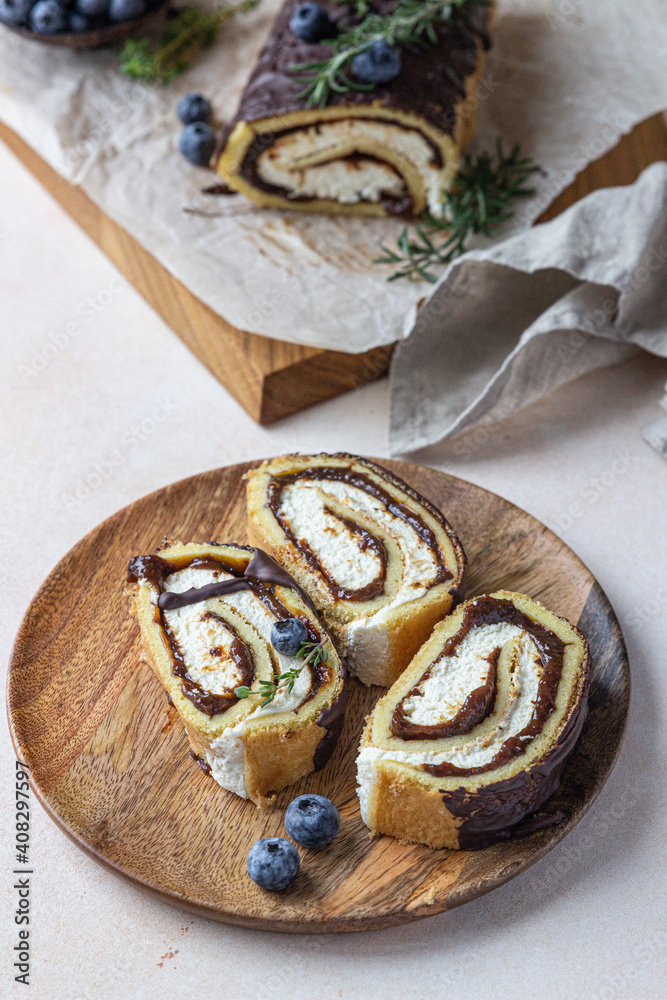 Sponge cake roll with chocolate and cream cheese decorated with chocolate glaze, blueberry and rosemary on parchment, light background. Biscuit swiss roll. Sponge cake roll with chocolate and cream cheese decorated with chocolate glaze, blueberry and rosemary on parchment, light background. Biscuit swiss roll.