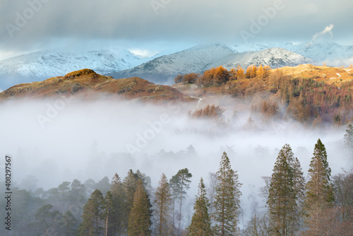Beautiful golden light illuminating Cumbrian fells on a Winter afternoon with mist filled valley and snowcapped mountains. Lake District, England, UK.