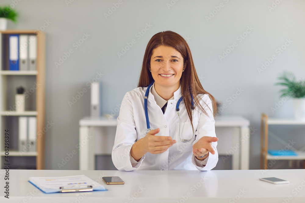 Smiling young woman doctor in white medical uniform sitting and gesticulating during online meeting with patient in medical clinic office. Telemedicine and online medicare concept Smiling young woman doctor in white medical uniform sitting and gesticulating during online meeting with patient in medical clinic office. Telemedicine and online medicare concept