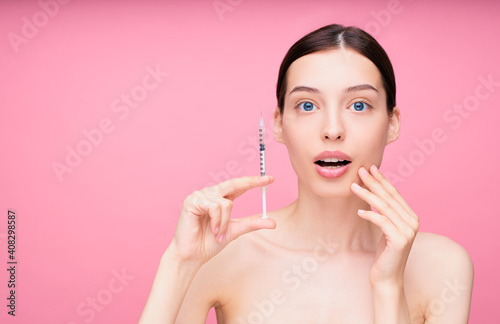 Surprised young girl model holds a syringe with botokos near her face.