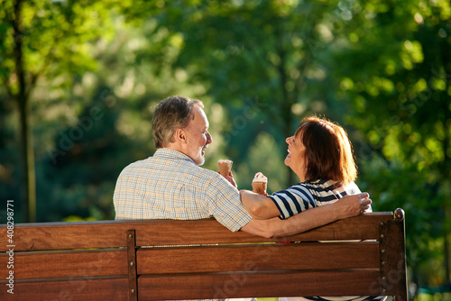 Happy retired couple eating ice-cream and laughing. Senior caucasian man and woman sit on the bench back view.