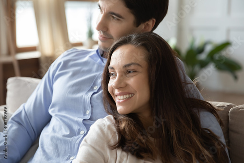 Close up dreamy smiling couple hugging, visualizing good future, looking to aside, sitting on cozy couch at home, happy man and woman cuddling, dreaming together, planning, enjoying lazy weekend