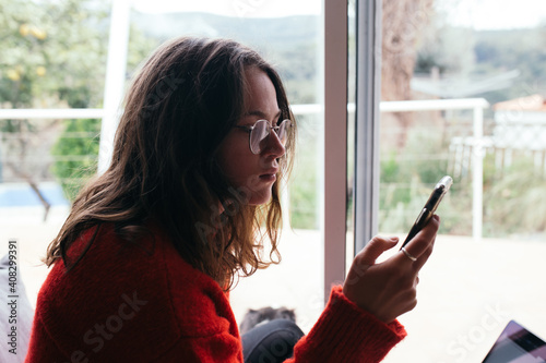 Attractive pretty young woman in prescription glasses look at phone screen. Beautiful soft light come from window, she wears cosy red wool sweater. Comfortable and calm weekend at home