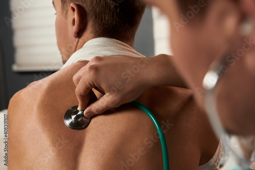doctor listens to the breath of a young white patient with a stethoscope on his back