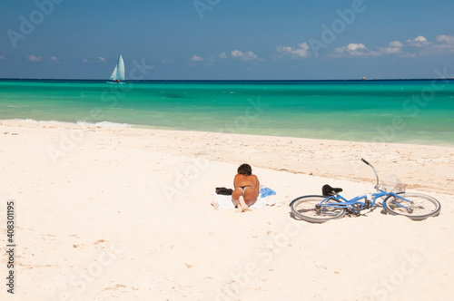 Fototapeta Naklejka Na Ścianę i Meble -  Young Caucasian woman unrecognizable, Tanning on a deserted Caribbean beach with his bicycle on the sand. On the horizon, the blue sky and a sailboat, Playa del Carmen, Mexico.