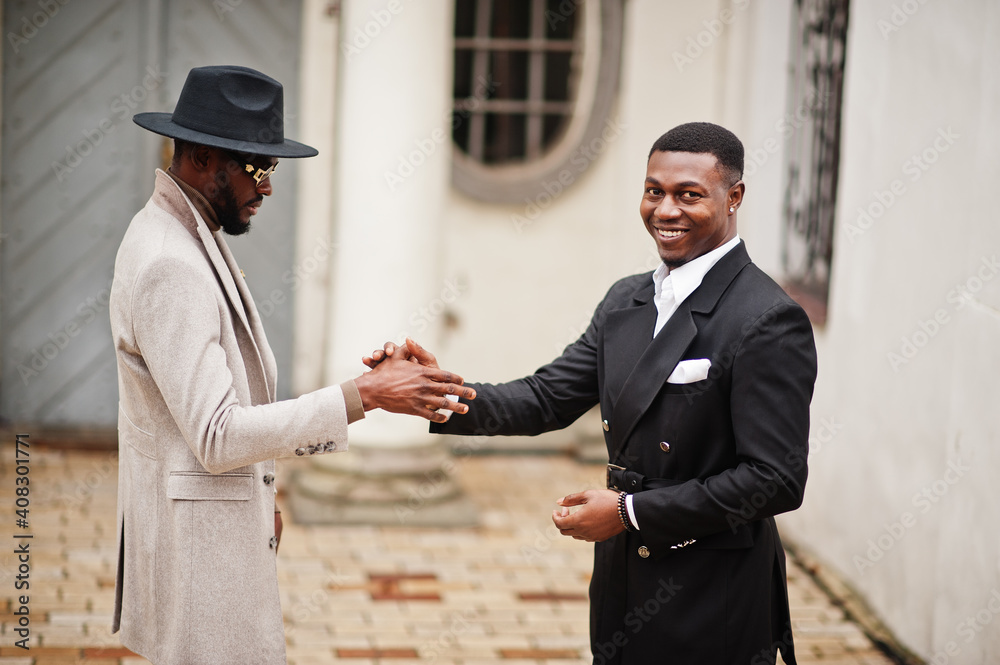 Two fashion black men shake hands each other. Fashionable portrait of african american male models. Wear suit, coat and hat.