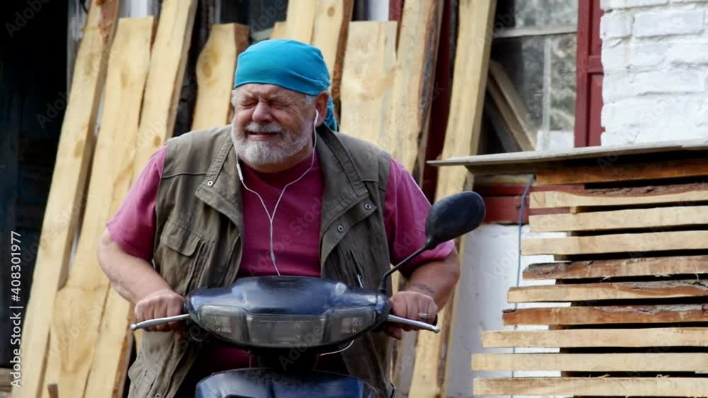 Caucasian bearded senior sitting on moped shows with expressive face and gesticulations how he would ride on it Caucasian bearded senior sitting on moped shows with expressive face and gesticulations how he would ride on it