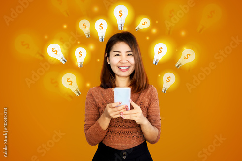 Billede på lærred smiling Asian woman holding mobile phone with lightbulb icon and dollar sign,  i