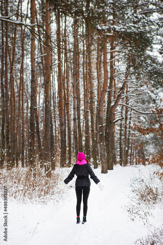 A girl in a pink scarf and hat stands in the middle of a pine forest. Beautiful pine forest on a frosty day.