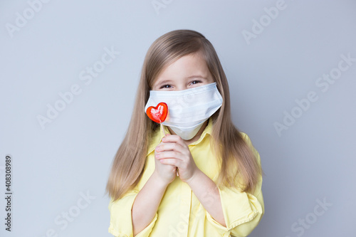 valentine's day caucasian child holding a lollipop heart over grey background. Face mask.Donation,heart health,world heart day, world health day,world mental health day.Health and heart concept.