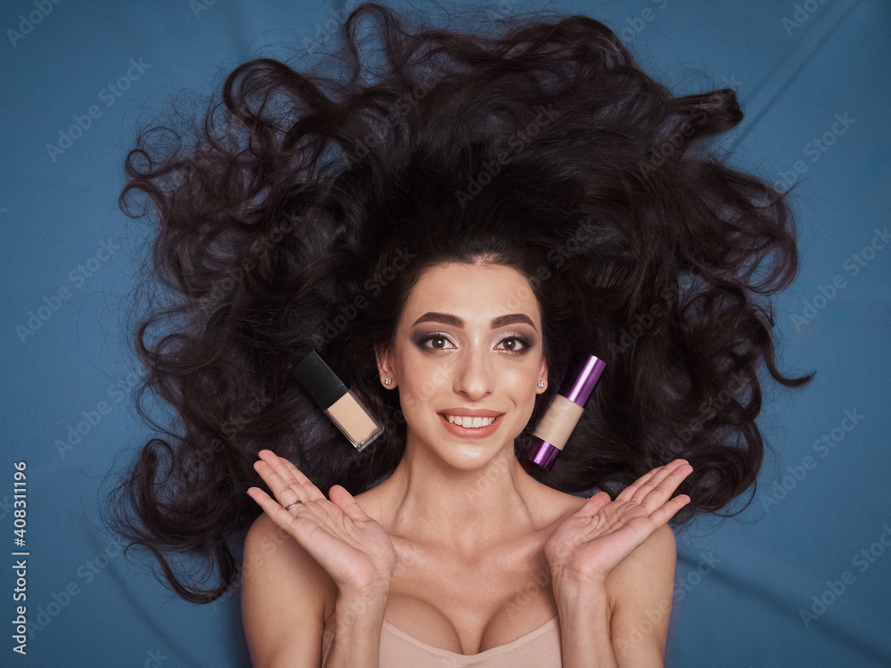 Fototapeta premium Skin care beautiful cosmetic makeup mixed race brunette model gorgeous hair full lips tan olive skin lying cobalt color backdrop professional cosmetic bottles and brushes