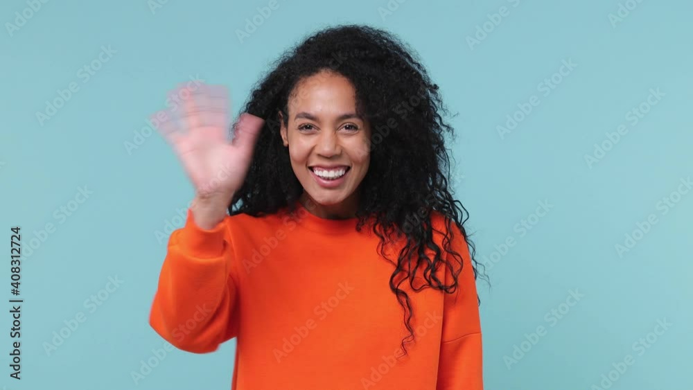 Smiling young african american woman curly hair 20s years old wearing orange sweatshirt waving meeting greeting with hand as notices someone isolated on pastel blue color background studio portrait